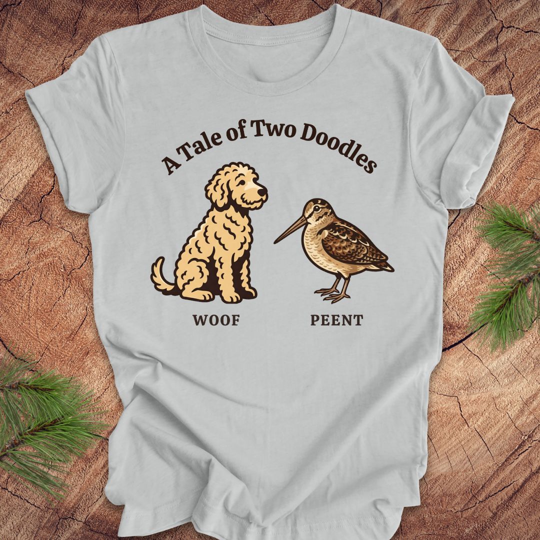 Gray t-shirt with a doodle dog and a woodcock bird with text, 'a tale of two doodles' on a wood background.