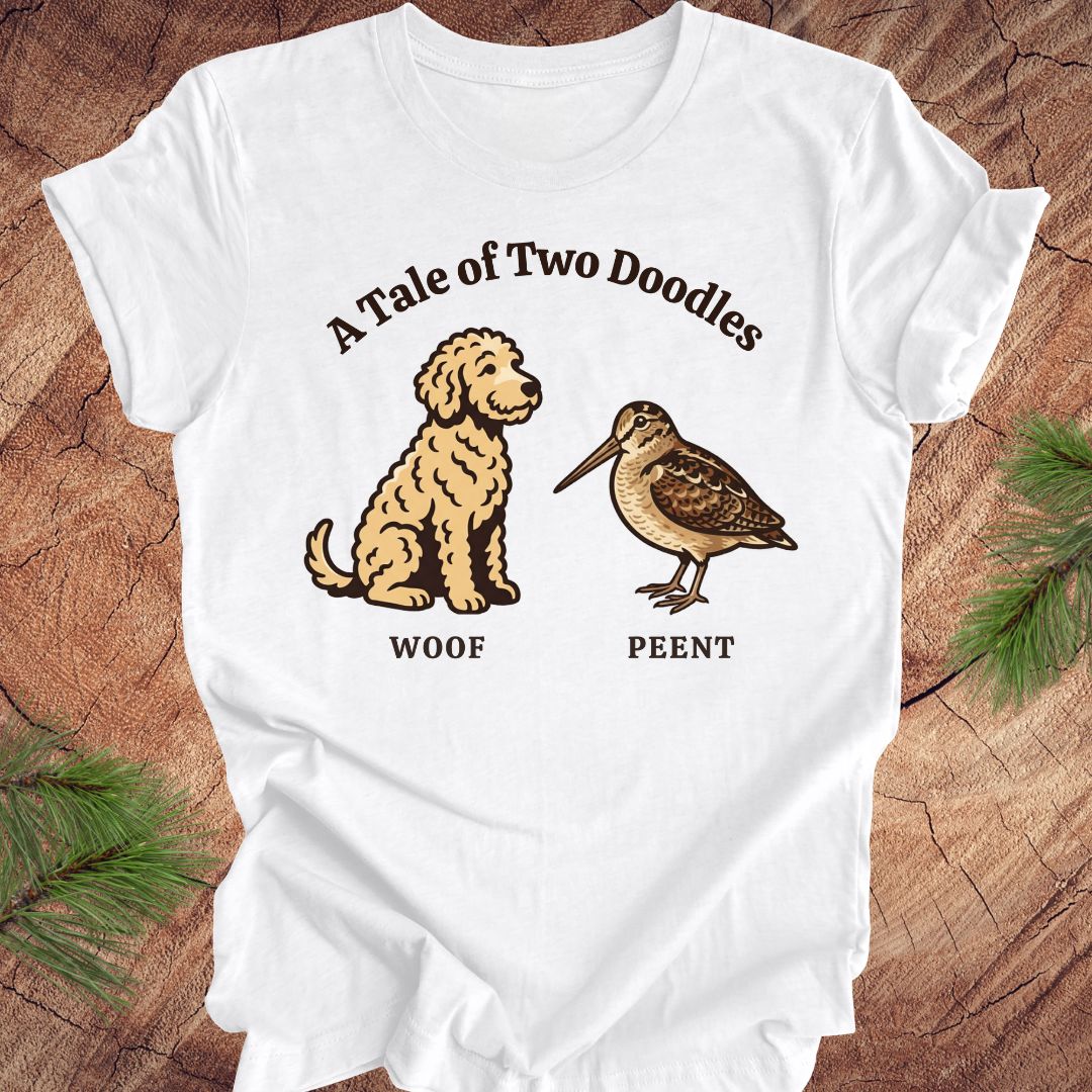 White t-shirt with a doodle dog and a woodcock bird with text, 'a tale of two doodles' on a wood background.
