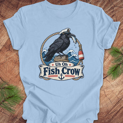 A light blue crewneck t-shirt featuring a detailed graphic of a black raven fish crow holding a silver fish, set against a nautical background with waves, ropes, and a banner that reads 'Uh Oh Fish Crow'. The t-shirt is displayed flat on a rustic wood background with pine needles in the corners.
