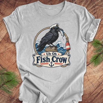 A light gray crewneck t-shirt laid flat on a rustic wood background with pine branches in the corners. The shirt features a detailed graphic of a black raven fish crow holding a red-and-white buoy on a rope, with a banner below reading 'Uh Oh Fish Crow' and an anchor.