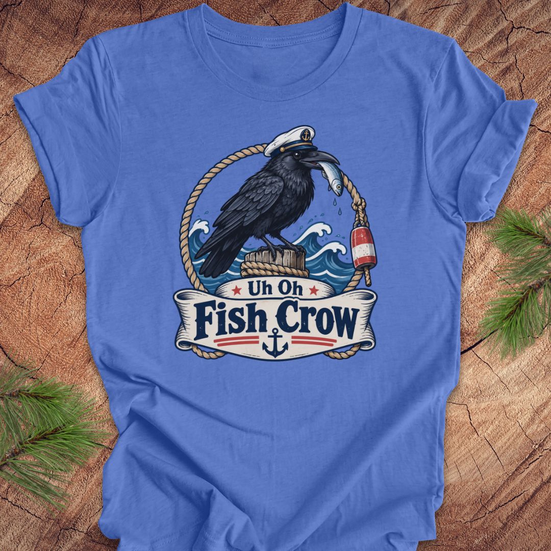 A blue short-sleeve crewneck t-shirt laid flat on a rustic wood background with pine branches in the corners. The shirt features a large graphic print of a black raven wearing a white captain's hat and holding a silver fish, with a banner below reading 'Uh Oh Fish Crow' and an anchor.