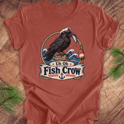 A rust-colored crewneck t-shirt featuring a detailed graphic of a black raven carrying a fish on its beak, framed by ropes and waves, with a banner below reading 'Uh Oh Fish Crow'. The shirt is laid flat on a rustic wood background with pine branches in the corners.