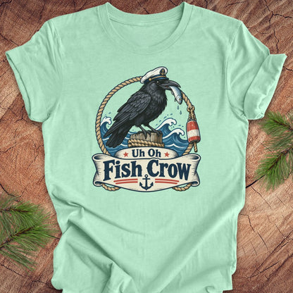 A mint green short-sleeve t-shirt with a detailed graphic of a black raven wearing a captain's hat and holding a silver fish, perched on a wooden post with ropes and waves. Below the illustration is a banner reading 'Uh Oh Fish Crow' with an anchor at the bottom.