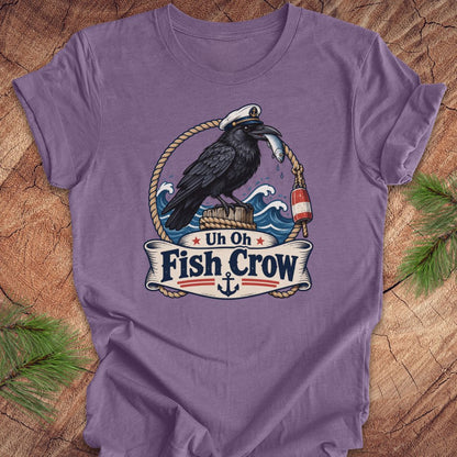 A purple short-sleeve t-shirt with a detailed graphic of a black raven crow holding a fish, set against a nautical background with waves, ropes, and a banner that reads 'Uh Oh Fish Crow'. The shirt is displayed flat on a rustic wood background with pine branches in the corners.