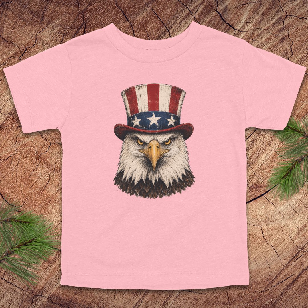 Pink toddler t-shirt with an eagle wearing a top hat on a wooden surface