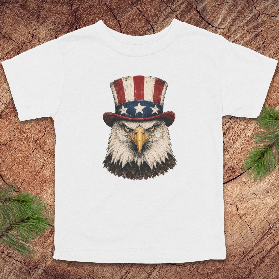 White toddler t-shirt with an eagle wearing a top hat on a wooden surface