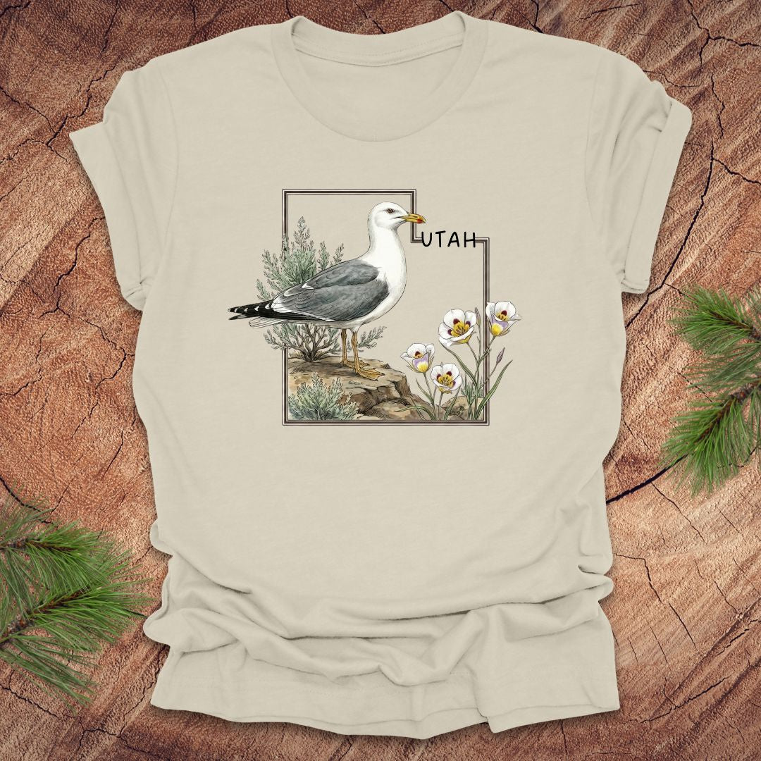 Beige t-shirt with an outline of the state of Utah and a California gull and Sego lily on a wooden surface