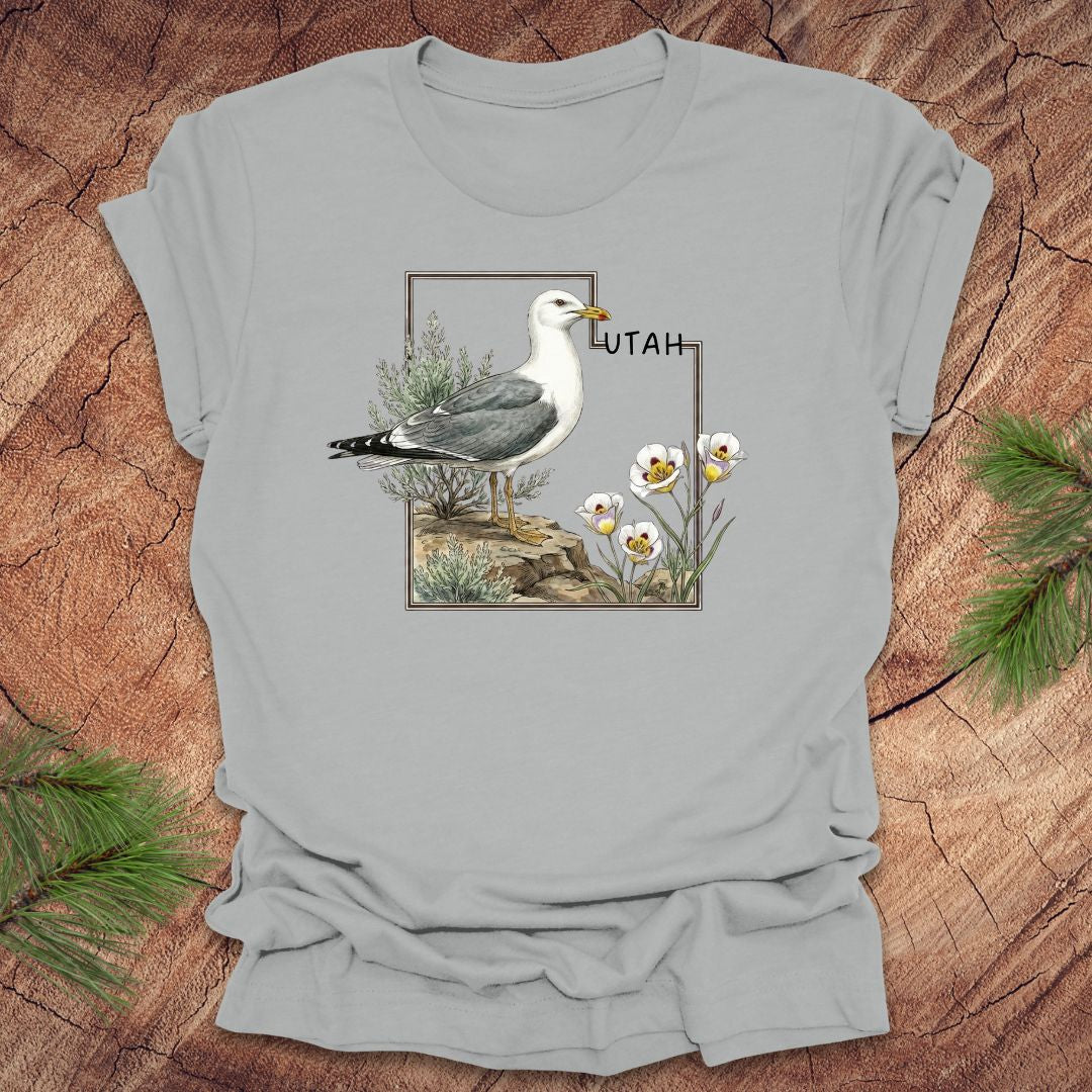 Silver t-shirt with an outline of the state of Utah and a California gull and Sego lily on a wooden surface