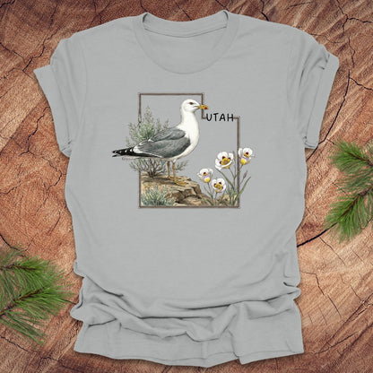 Silver t-shirt with an outline of the state of Utah and a California gull and Sego lily on a wooden surface