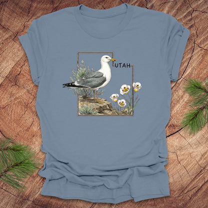 Steel blue t-shirt with an outline of the state of Utah and a California gull and Sego lily on a wooden surface