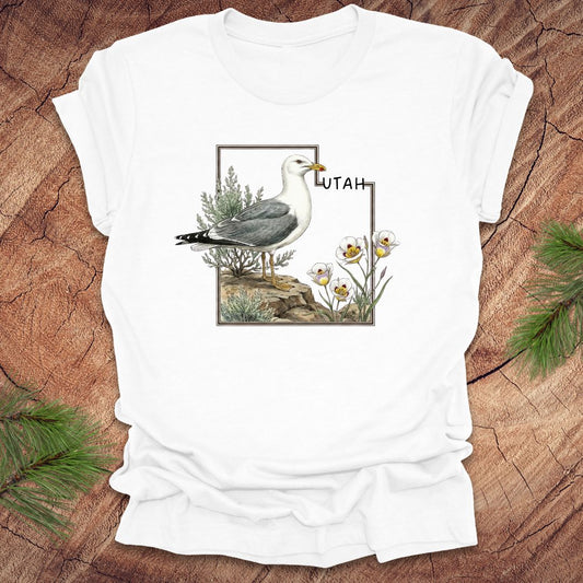 White t-shirt with an outline of the state of Utah and a California gull and Sego lily on a wooden surface