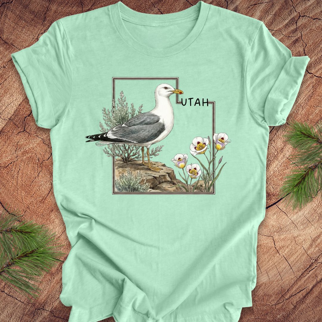 Mint green t-shirt with an outline of the state of Utah and a California gull and Sego lily on a wooden surface