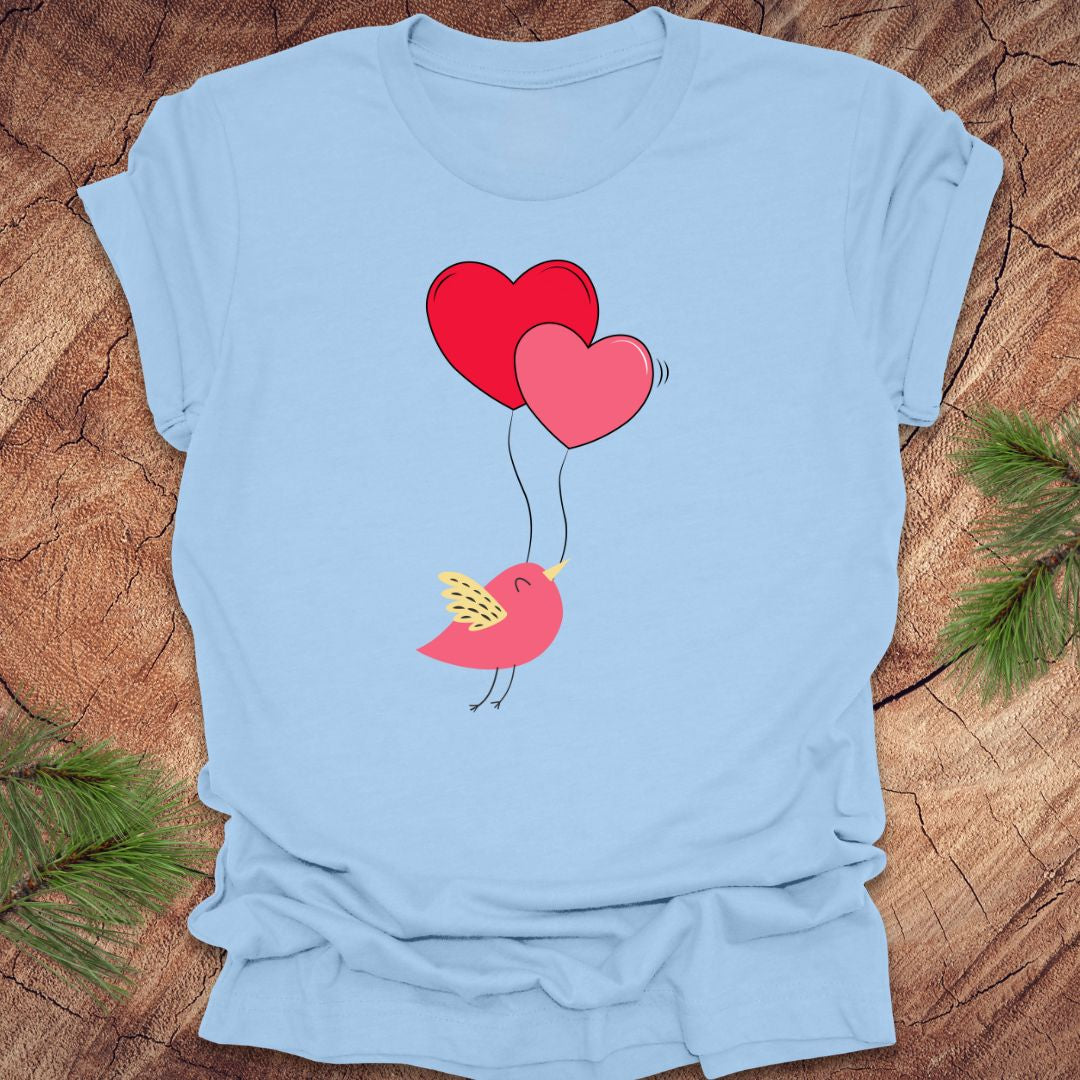 Baby blue t-shirt with a cartoon bird on the strings of 2 heart-shaped balloons, on a wooden background.