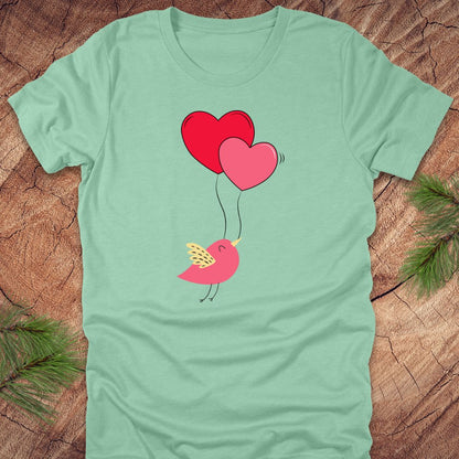 Mint green t-shirt with a cartoon bird on the strings of 2 heart-shaped balloons, on a wooden background.