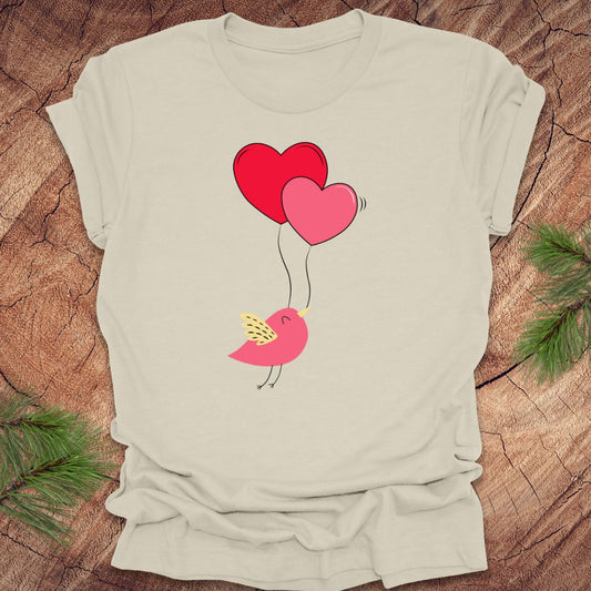 Beige t-shirt with a cartoon bird on the strings of 2 heart-shaped balloons, on a wooden background.