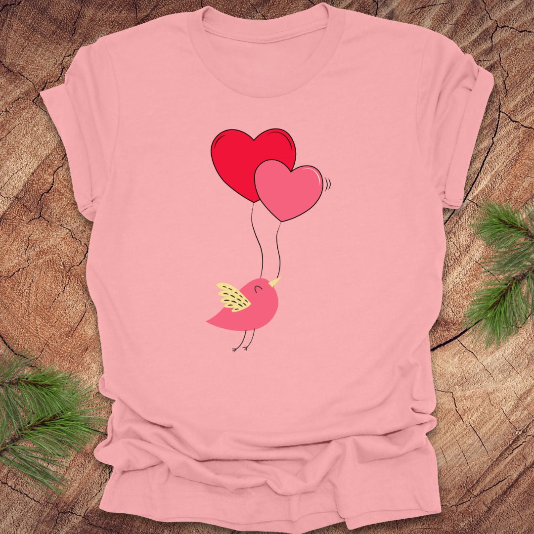 Pink t-shirt with a cartoon bird on the strings of 2 heart-shaped balloons, on a wooden background.