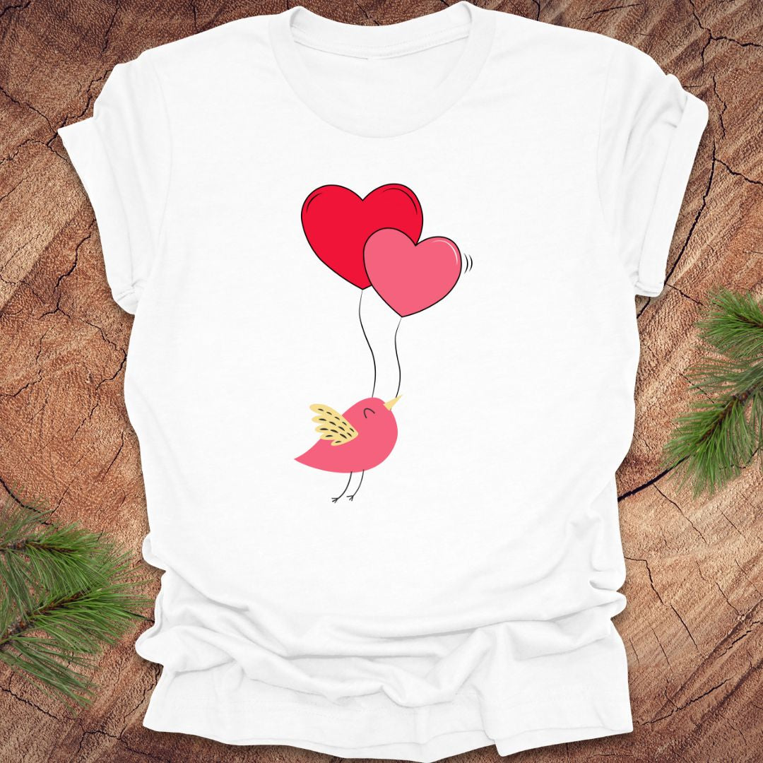 White t-shirt with a cartoon bird on the strings of 2 heart-shaped balloons, on a wooden background.