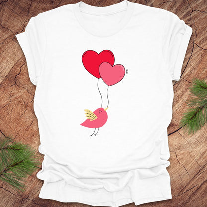 White t-shirt with a cartoon bird on the strings of 2 heart-shaped balloons, on a wooden background.