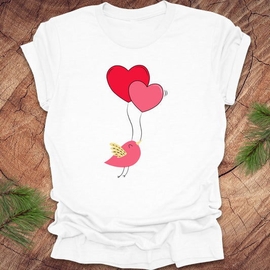 White t-shirt with a cartoon bird on the strings of 2 heart-shaped balloons, on a wooden background.