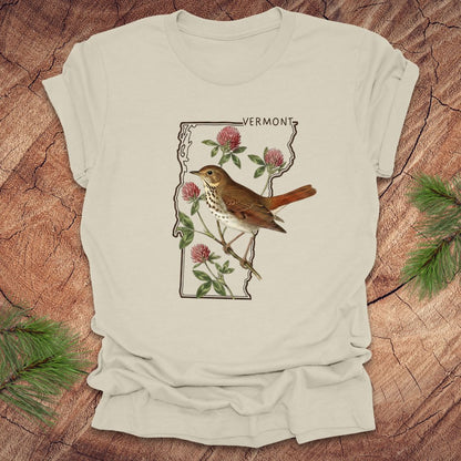 Beige t-shirt with a graphic of Vermont, a bird, and flowers on a wooden surface.