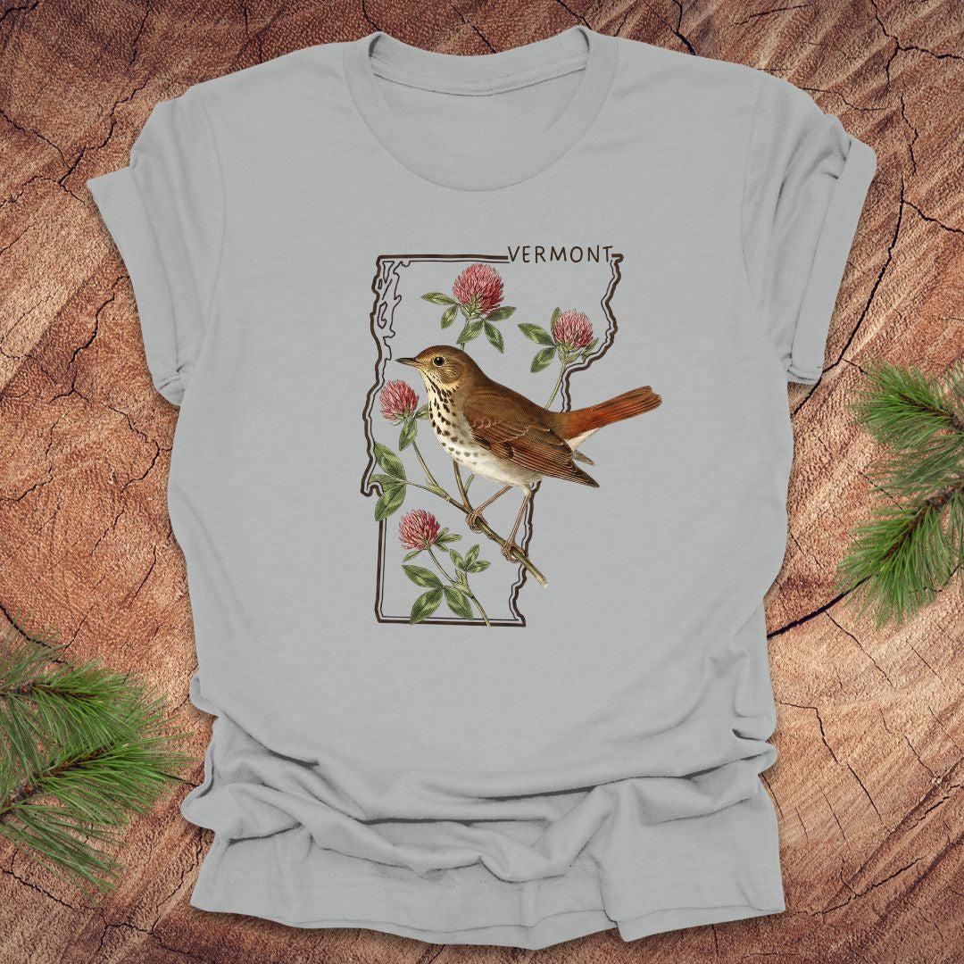 Light gray t-shirt with a bird and floral design on Vermont's outline on a wooden surface.