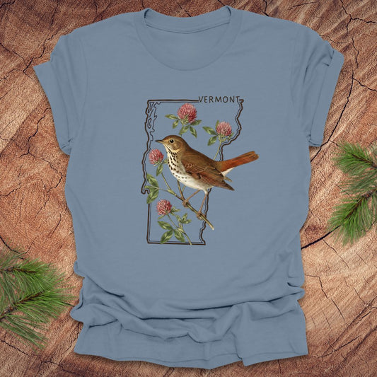 Blue t-shirt with a bird and floral design on Vermont's outline, placed on a wooden surface.