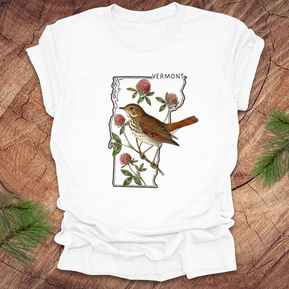White t-shirt with Vermont state design featuring a bird and flowers on a wooden surface.