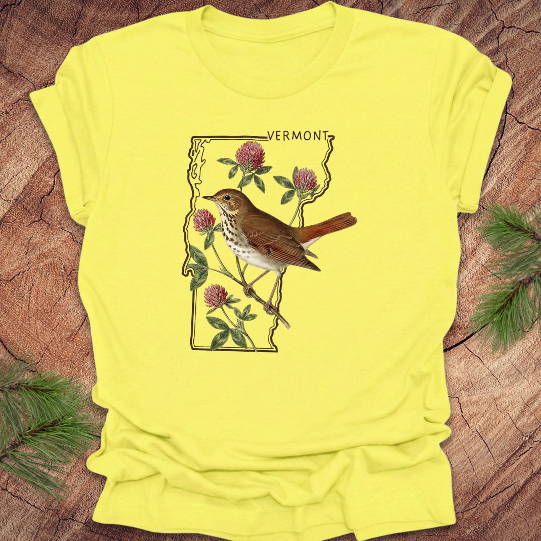 Yellow t-shirt with a bird and floral design on Vermont's outline, placed on a wooden surface.