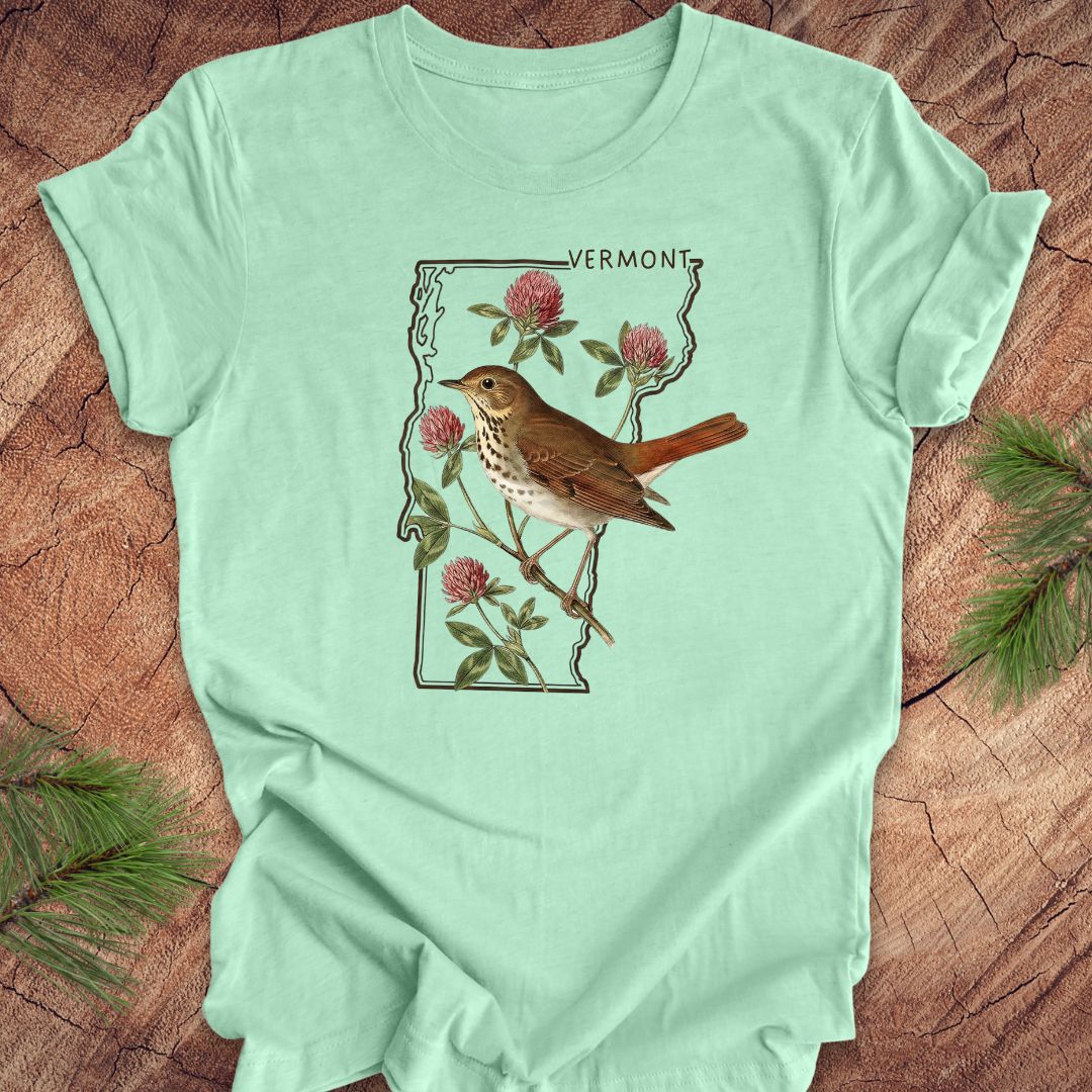 Mint green t-shirt with a bird and floral design on Vermont's outline, placed on a wooden surface.