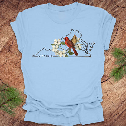 Light blue t-shirt with a map of Virginia featuring cardinals and flowers on a wooden surface.