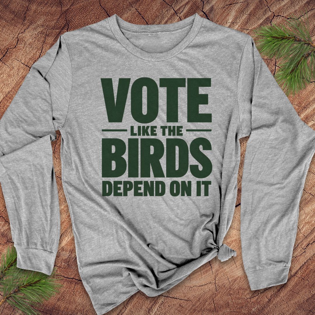 A heather gray long-sleeve t-shirt with a dark green graphic text reading 'VOTE LIKE THE BIRDS DEPEND ON IT', laid flat on a rustic wood background with pine branches in the corners.