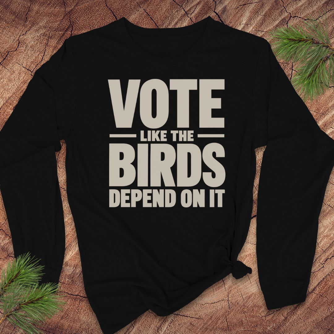 A black long-sleeve t-shirt with a bold beige graphic print reading 'VOTE LIKE THE BIRDS DEPEND ON IT', styled flat on a rustic wood background with pine needles.