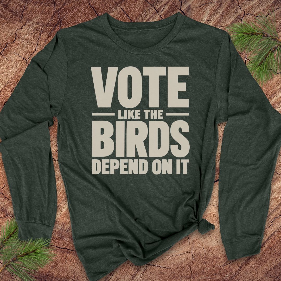 A dark forest green long-sleeve t-shirt with a bold cream-colored graphic text reading 'VOTE LIKE THE BIRDS DEPEND ON IT'. The shirt is styled flat on a textured brown wood background with pine needles in the corners.