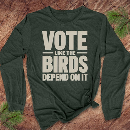 A dark forest green long-sleeve t-shirt with a bold cream-colored graphic text reading 'VOTE LIKE THE BIRDS DEPEND ON IT'. The shirt is styled flat on a textured brown wood background with pine needles in the corners.