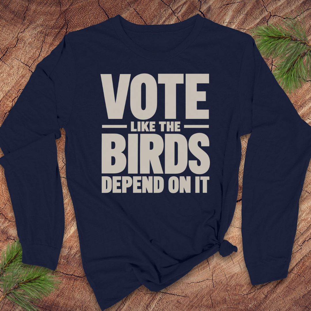 A long-sleeve navy blue t-shirt with a beige graphic print that reads 'VOTE LIKE THE BIRDS DEPEND ON IT', styled on a rustic wood background with pine needles.