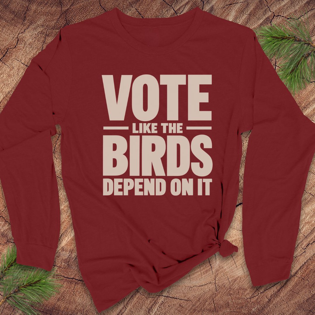 A long-sleeve maroon t-shirt with a beige graphic print that reads 'VOTE LIKE THE BIRDS DEPEND ON IT', styled on a rustic wood background with pine needles.