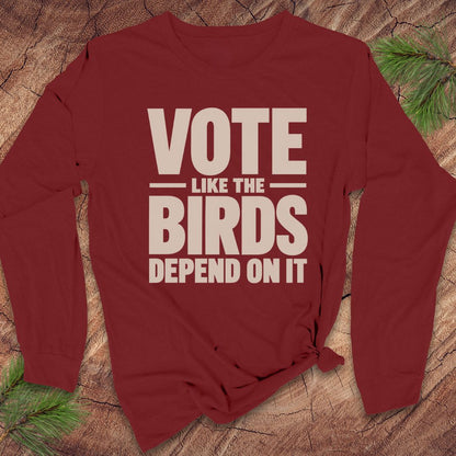 A long-sleeve maroon t-shirt with a beige graphic print that reads 'VOTE LIKE THE BIRDS DEPEND ON IT', styled on a rustic wood background with pine needles.