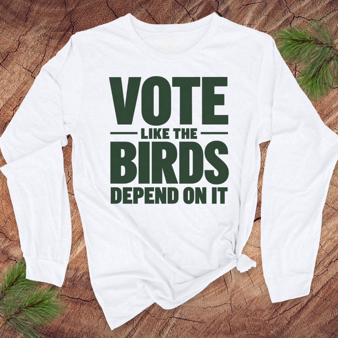 A long-sleeve white t-shirt with a beige graphic print that reads 'VOTE LIKE THE BIRDS DEPEND ON IT', styled on a rustic wood background with pine needles.