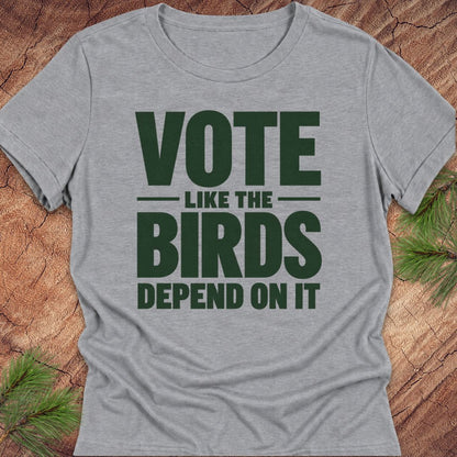A heather gray short-sleeve t-shirt with a dark green printed slogan reading 'VOTE LIKE THE BIRDS DEPEND ON IT', laid flat on a rustic wood surface with pine branches in the corners.