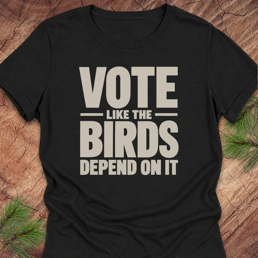 A black short-sleeve t-shirt with a bold beige graphic print reading 'VOTE LIKE THE BIRDS DEPEND ON IT', laid flat on a rustic wood background with pine branches in the corners.