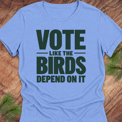 A light blue short-sleeve t-shirt with a dark green graphic text reading 'VOTE LIKE THE BIRDS DEPEND ON IT', laid flat on a rustic wood background with pine needles in the corners.