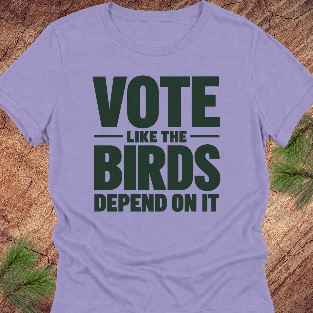 A lavender short-sleeve crewneck t-shirt with a dark green graphic text reading 'VOTE LIKE THE BIRDS DEPEND ON IT', displayed flat on a rustic wood background with pine needles.