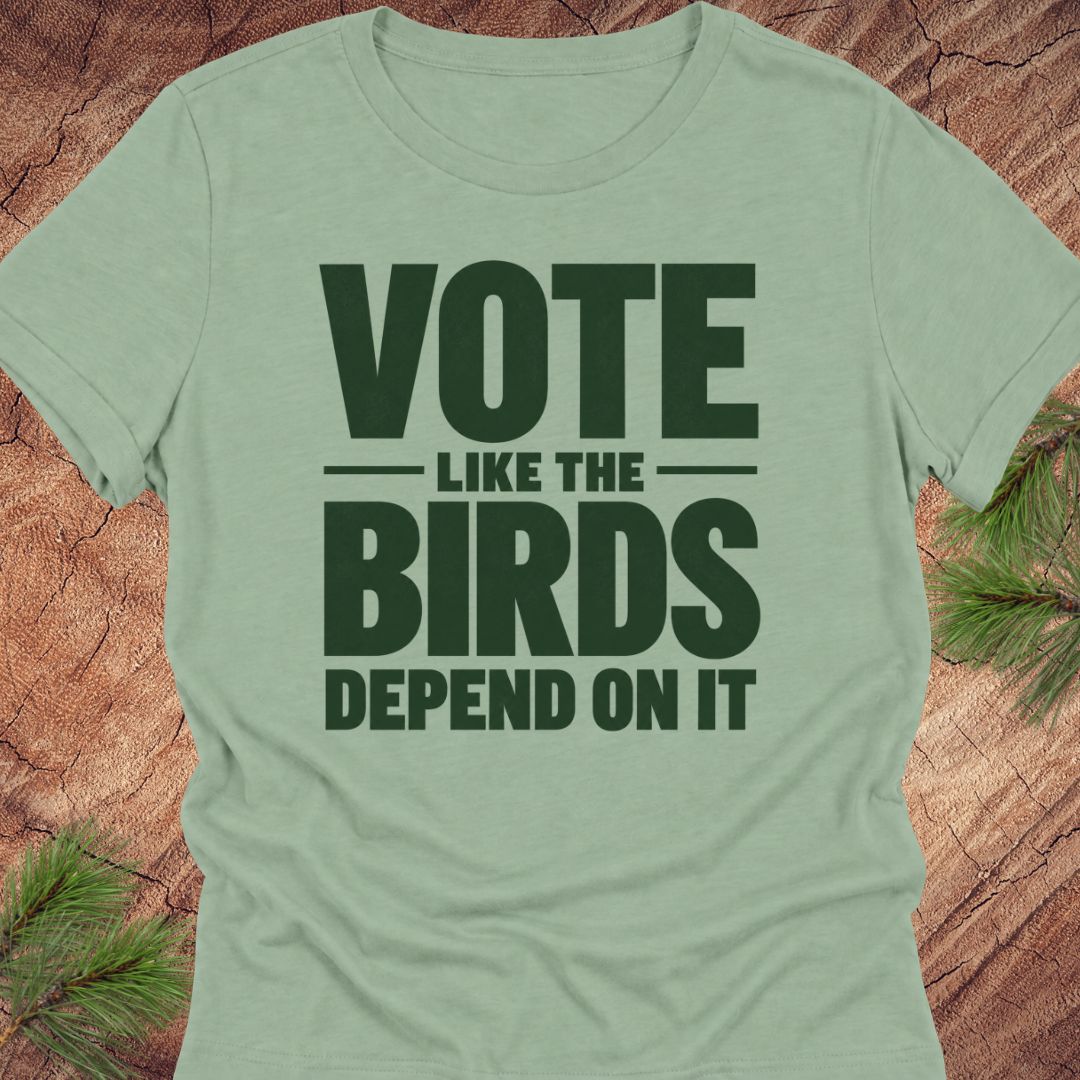 A sage green short-sleeve t-shirt with a dark green graphic text reading 'VOTE LIKE THE BIRDS DEPEND ON IT', displayed flat on a rustic wood background with pine needles in the corners.