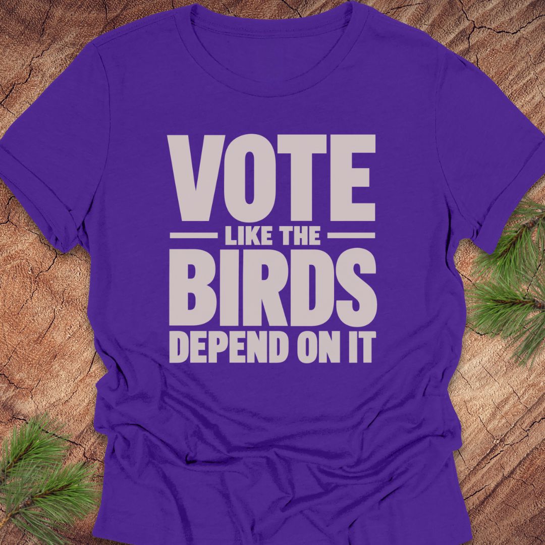 A purple short-sleeve t-shirt with the slogan 'VOTE LIKE THE BIRDS DEPEND ON IT' printed in bold white text, laid flat on a rustic wood background with pine branches in the corners.
