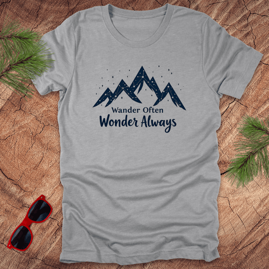 A gray t-shirt with the text 'Wander Often Wonder Always' printed on the front, featuring a mountain graphic. The shirt is displayed on a wooden surface with pine needles and a pair of sunglasses.