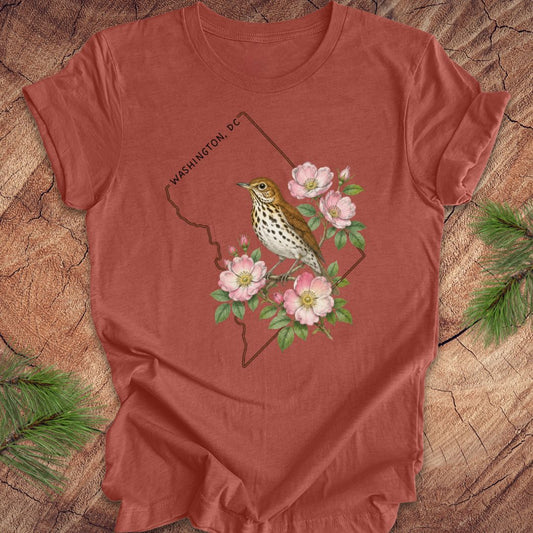 Rust colored t-shirt with a Washington DC outline and  thrush and rose design on a wooden surface
