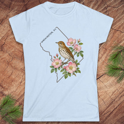 A light blue short-sleeve crewneck t-shirt featuring a graphic of a brown bird perched on pink flowers with green leaves, set against a map outline labeled 'WASHINGTON, DC'. The shirt is displayed flat on a rustic wood background with pine needles.