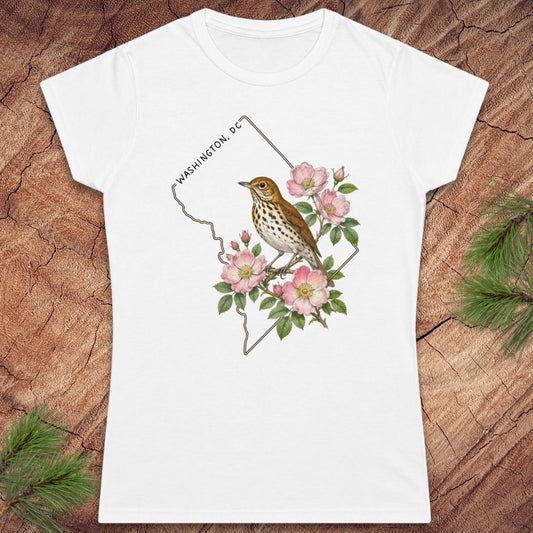 A white short-sleeve crewneck t-shirt featuring a graphic print of a bird perched on pink flowers with the text 'WASHINGTON, DC' inside a thin outline shape. The t-shirt is displayed flat on a rustic wood background with pine needles in the corners.