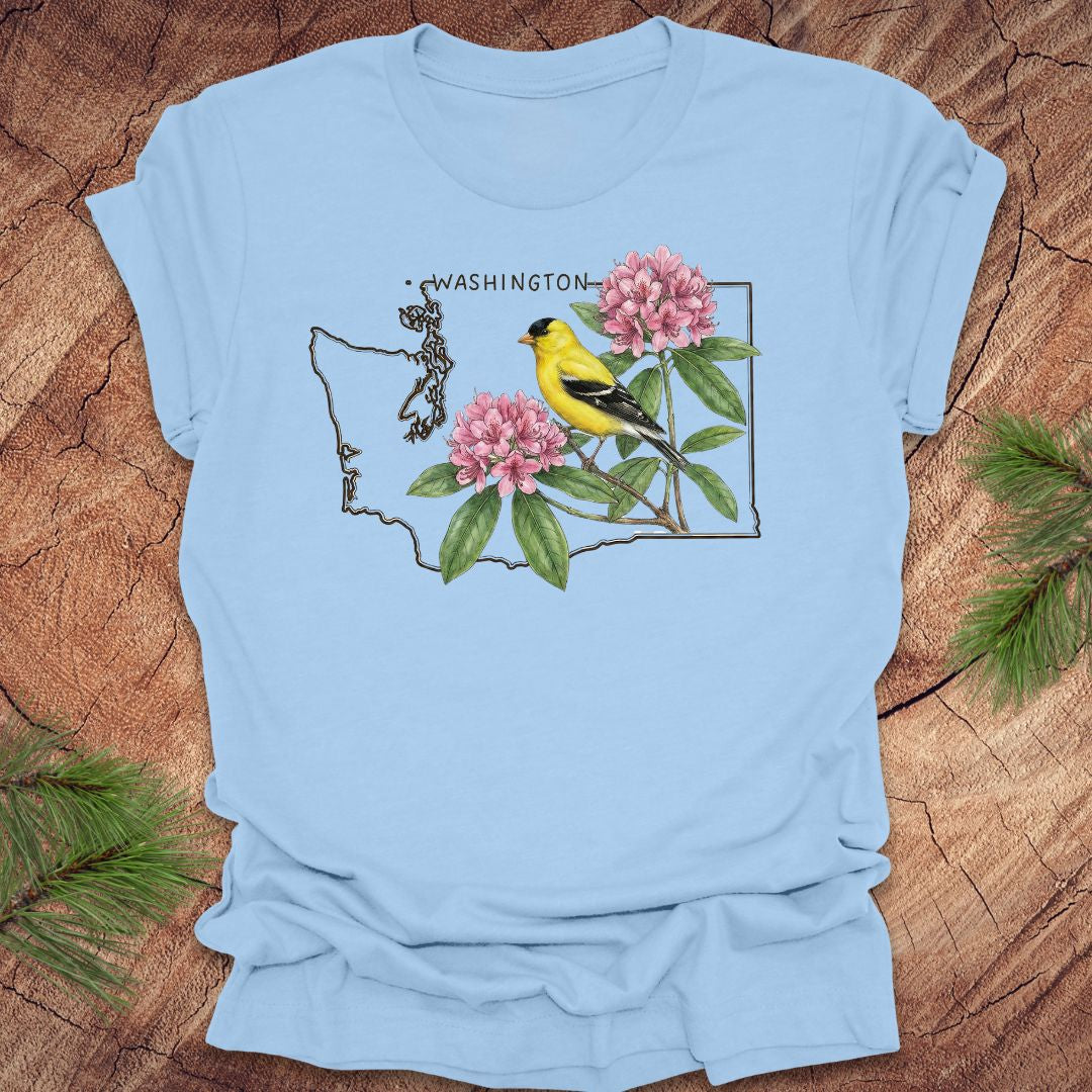 Baby blue t-shirt with Washington state outline with state bird goldfinch and rhododendron on a wood background.