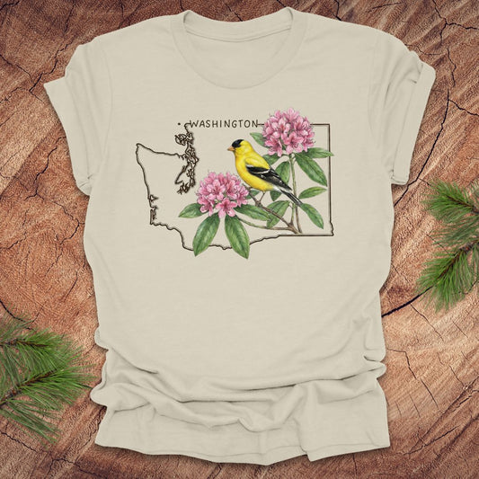 Beige t-shirt with Washington state outline with state bird goldfinch and rhododendron on a wood background.